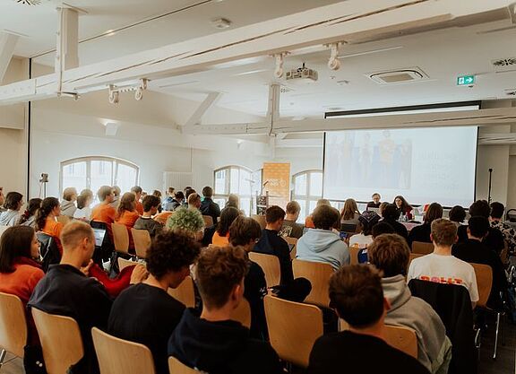Junge Menschen sitzen auf Stühlen in einem große Saal und schauen nach vorn auf eine Leinwand.